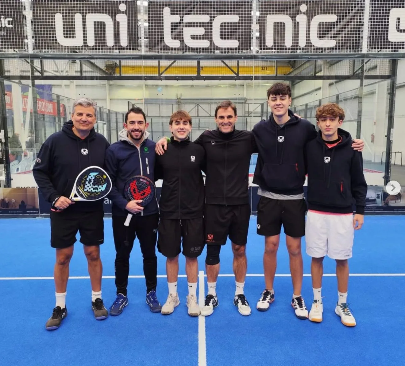 Grupo masculino de jugadores y monitores posando con palas en pista indoor de Pádel Pista Norte, con equipación del club y sponsor uni tecnic